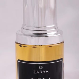 Könnte beinhalten: Eine klare Glasflasche mit silbernem Verschluss und schwarzem Etikett. Die Flasche enthält eine goldene Flüssigkeit mit kleinen Partikeln. Auf dem Etikett steht "ZARYA 4 Liebe MAGISCHES RITUALOL".