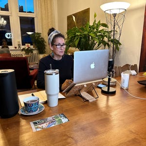 Puede incluir: Una persona está sentada en una mesa de madera con un portátil, un vaso blanco y una taza de té. Una tetera negra y una pequeña tarjeta también están sobre la mesa. La habitación tiene un ambiente cálido, con una ventana grande y una lámpara decorativa.