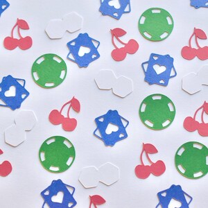 Op de afbeelding: Een verzameling papieren confetti-vormen, waaronder groene ronde pokerchips, rode dubbele kersen, blauwe vierkante speelkaarten met hartvormige uitsparingen en witte zeshoekige stukjes verspreid over een wit oppervlak.