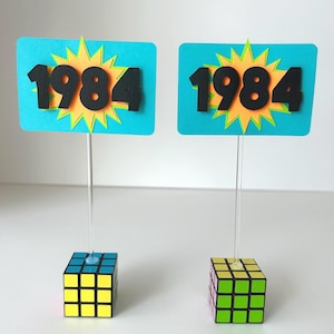 Op de afbeelding: Twee blauwe en turquoise kartonnen borden met een geel en oranje stervormig ontwerp en het getal 1984 in het zwart. De borden zijn bevestigd aan heldere plastic stokjes die in een zwarte, witte, gele, groene en roze Rubik's Cube worden gestoken.
