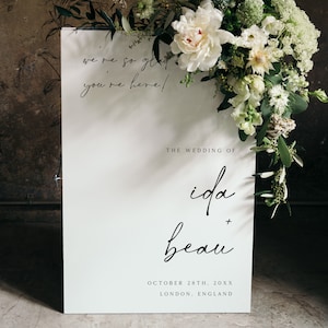 May include: White wedding sign with black calligraphy text that reads "We're so glad you're here!"  The wedding of Ida + Beau. October 28th, 20XX. London, England.
