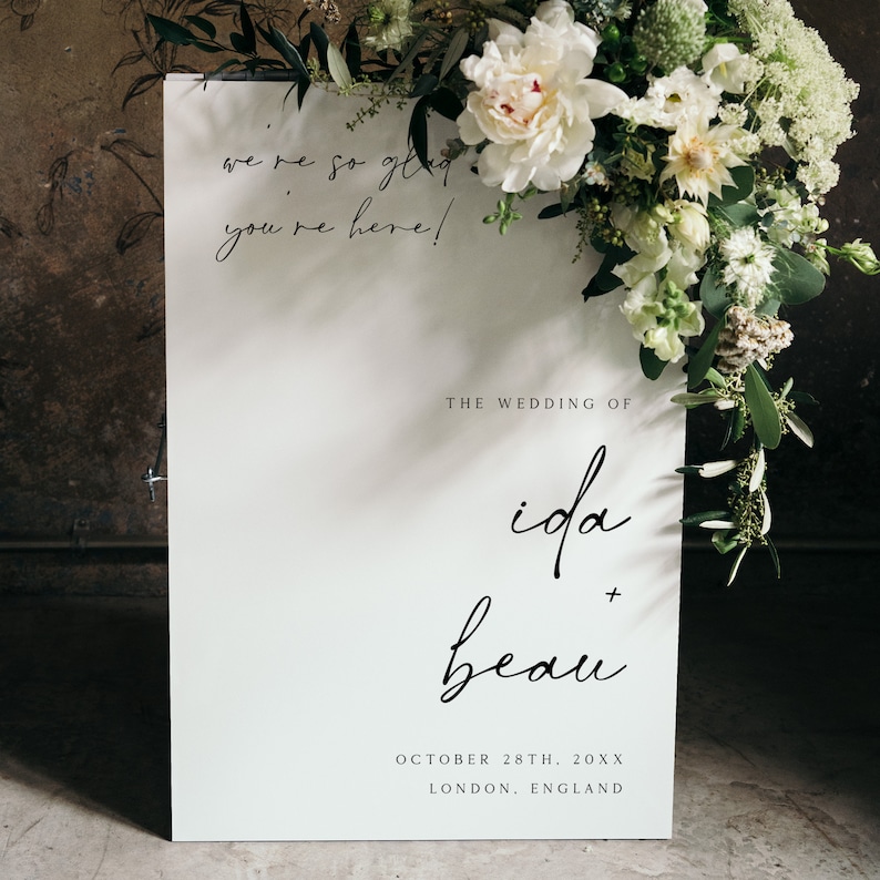May include: White wedding sign with black calligraphy text that reads "We're so glad you're here!"  The wedding of Ida + Beau. October 28th, 20XX. London, England.