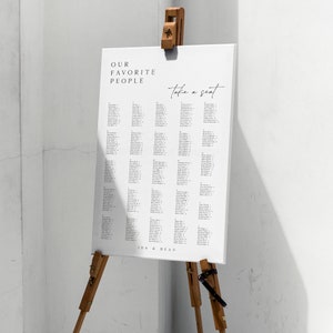 May include: A white seating chart with the text "Our Favorite People" and "Take a Seat" written in black. The chart lists names and table numbers. The chart is displayed on a wooden easel with the text "Ida & Beau" at the bottom.