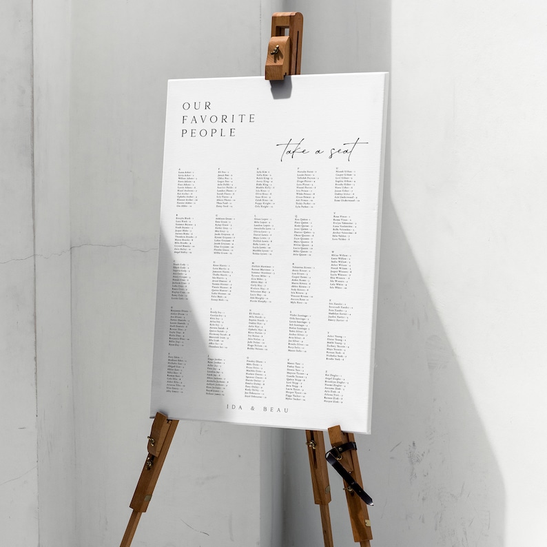 May include: A white seating chart with the text "Our Favorite People" and "Take a Seat" written in black. The chart lists names and table numbers. The chart is displayed on a wooden easel with the text "Ida & Beau" at the bottom.
