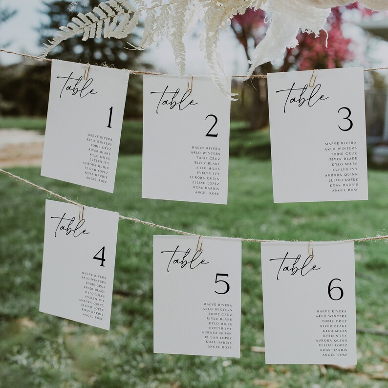 May include: Six white cards with black text, hanging from a string. Each card has a table number from 1 to 6, and a list of names. The cards are on a green background.