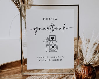 May include: A white sign in a gold frame with the text "PHOTO guestbook" and a black and white illustration of an instant camera. The text below the image reads "SNAP IT, SHAKE IT, STICK IT, SIGN IT."