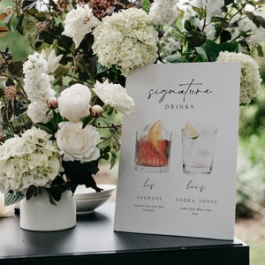 May include: A white sign with black text that reads "signature drinks". The sign features two watercolor illustrations of cocktails, one labeled "his negroni" and the other labeled "hers vodka tonic". The sign is sitting on a black table with white flowers in a vase.