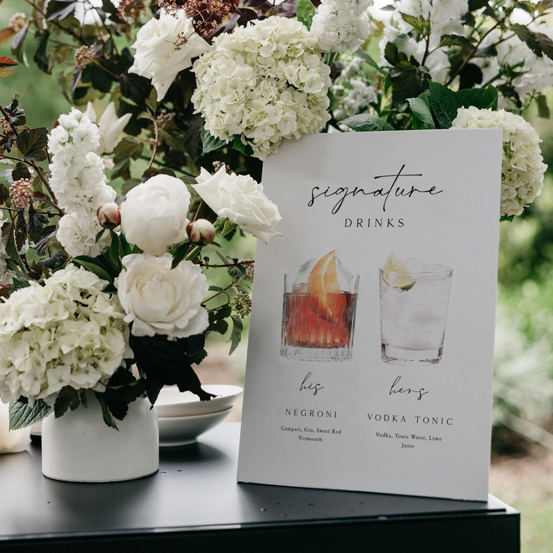 May include: A white sign with black text that reads "signature drinks". The sign features two watercolor illustrations of cocktails, one labeled "his negroni" and the other labeled "hers vodka tonic". The sign is sitting on a black table with white flowers in a vase.