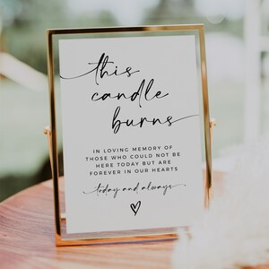 May include: A white sign in a gold frame with the text "this candle burns" in black cursive writing. The sign also includes the text "In loving memory of those who could not be here today but are forever in our hearts today and always" with a heart symbol.