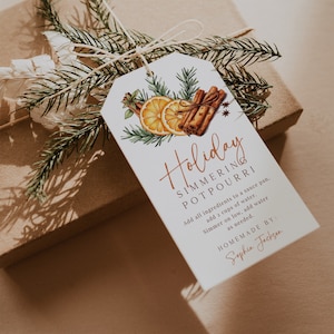 Könnte beinhalten: Ein weißer Geschenkanhänger mit einer handschriftlichen Aufschrift für ein "Weihnachtliches Potpourri zum Köcheln". Der Anhänger zeigt eine Aquarell-Illustration von Orangen, Zimtstangen und Grünzeug. Der Text lautet: "Alle Zutaten in einen Topf geben, 2 Tassen Wasser hinzufügen. Bei schwacher Hitze köcheln lassen, bei Bedarf Wasser hinzufügen. Selbstgemacht von: Sophia Jackson."