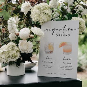 May include: A white sign with black text that reads "signature drinks" with illustrations of two cocktails. The text below the illustrations reads "his gin & tonic gin, tonic water, lime juice" and "hers paloma grapefruit juice, tequila, simple syrup, sparkling water". The bottom of the sign reads "Lottie Tommy | 28.10.xx".