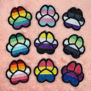 Op de afbeelding: Negen geborduurde pootafdrukpatches met verschillende regenboog- en pride-vlagkleurenschema's. Elke patch heeft een zwarte rand en is geplaatst tegen een roze achtergrond. De patches zijn gerangschikt in een raster van drie rijen en drie kolommen.