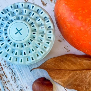 Puede incluir: Una bola de lavado azul claro con un dise&ntilde;o circular y una "X" blanca en el centro. La bola est&aacute; sobre una superficie de madera blanca con una hoja marr&oacute;n y una peque&ntilde;a nuez marr&oacute;n.