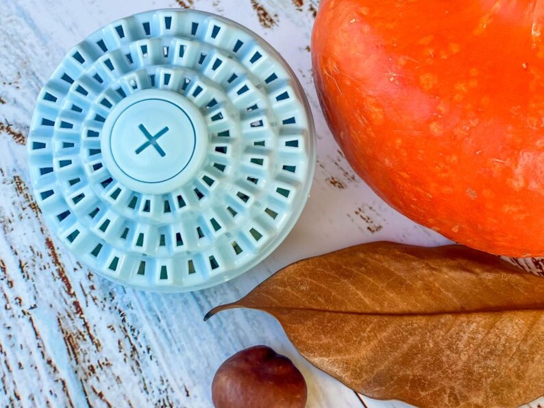 Puede incluir: Una bola de lavado azul claro con un dise&ntilde;o circular y una "X" blanca en el centro. La bola est&aacute; sobre una superficie de madera blanca con una hoja marr&oacute;n y una peque&ntilde;a nuez marr&oacute;n.