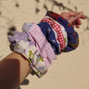 Könnte beinhalten: Eine Sammlung bunter Haargummis in verschiedenen Mustern, darunter Blumen-, Vichy- und nautische Designs. Die Haargummis sind aus Stoff und werden zusammengehalten, wodurch eine Vielzahl von Farben und Texturen gezeigt wird.