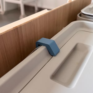 Könnte beinhalten: Ein hellblauer Kunststoffclip, der ein helles Tablett an einer Holzoberfläche befestigt. Das Tablett ist cremefarben und hat eine rechteckige Form. Die Holzoberfläche ist hellbraun.