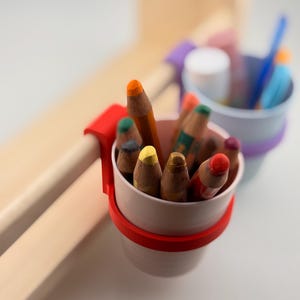 Könnte beinhalten: Nahaufnahme eines weißen und roten Stifthalters, gefüllt mit Buntstiften. Der Halter ist an einer hellen Holzoberfläche befestigt. Ein weiterer Stifthalter ist im Hintergrund zu sehen, mit einem lila und weißen Band.