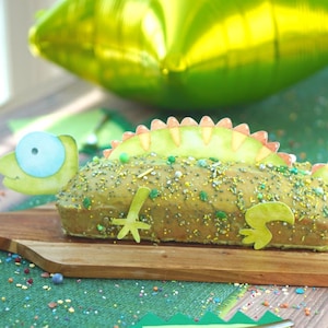 Könnte beinhalten: Ein Kuchen, der wie ein grünes Chamäleon dekoriert ist, komplett mit Kopf, Schwanz und Rückenstacheln. Der Kuchen liegt auf einem hölzernen Schneidebrett, ist mit bunten Konfetti bestreut und vor einem grünen und braunen Hintergrund platziert.