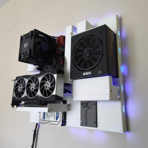 Könnte beinhalten: Ein wandmontiertes Open-Frame-Computergehäuse mit weißer Rückplatte und blauer LED-Kantenbeleuchtung. Zu den Komponenten gehören ein schwarzer CPU-Kühler, eine Grafikkarte mit drei Lüftern, ein 650-Watt-Netzteil, eine Festplatte und eine Kingston-SSD.