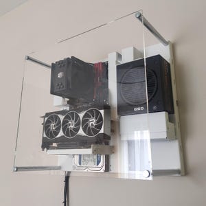 May include: A wall-mounted computer case featuring an open-frame design with a clear acrylic front panel held by four silver metal standoffs. The internal components include a black CPU cooler, a triple-fan graphics card, and a black power supply.