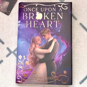 Può includere: Un libro e un segnalibro con il titolo "Once Upon a Broken Heart" e un'illustrazione romantica di una coppia. La copertina del libro è scura con dettagli dorati e il nome dell'autore, Stephanie Garber, è visibile.