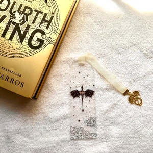 Peut inclure: Un marque-page transparent avec un motif de dragon noir et un ruban blanc. Le marque-page est doté d'un charme de dragon doré attaché au ruban. Le marque-page est posé sur une surface blanche à côté d'un livre doré avec le titre "Fourth Wing" sur la couverture.