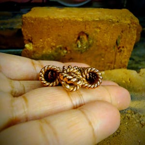 Puede incluir: Primer plano de una mano sosteniendo un conjunto de cuatro anillos de color dorado, con forma de cuerda. Los anillos están trenzados y anudados entre sí.