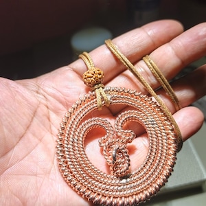 Puede incluir: Collar con colgante de color cobre con diseño de corazón. El colgante está hecho de alambre enrollado y cuelga de un cordón marrón. El collar se sostiene en una mano, mostrando la intrincada artesanía y los tonos cálidos del metal.