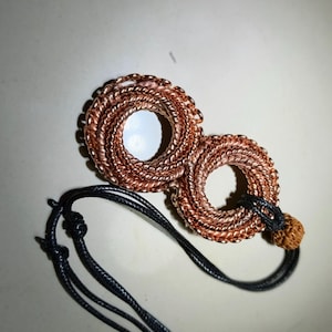 Puede incluir: Un collar con colgante de color cobre con dos diseños circulares entrelazados. Los círculos están hechos de alambre enrollado, con un cordón negro y una pequeña cuenta redonda marrón.