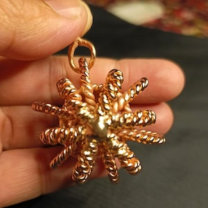 Peut inclure: Un pendentif doré avec un motif de soleil. Le pendentif présente une sphère centrale avec des rayons en fil torsadé. Le pendentif est maintenu par une petite boucle en haut.