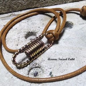 May include: A brown cord necklace with a copper-colored pendant. The pendant is made of a series of stacked rings and has a loop at the top. The text "Acuvac Sacred Cubit" is visible in the image.