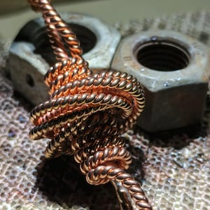 Op de afbeelding: Close-up van een koperkleurig, gedraaid metalen sieraad. Het ontwerp heeft een geknoopt gedeelte en een gevlochten touwachtige structuur. De achtergrond bevat metalen moeren en een getextureerd oppervlak.