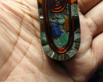 Può includere: Un ciondolo ovale trasparente con una spirale di filo di rame all'interno. Il ciondolo è pieno di piccole pietre preziose colorate, tra cui verde, blu e arancione.