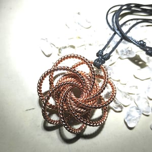 Puede incluir: Un collar con colgante de color cobre con un diseño circular tejido. El colgante está suspendido de un cordón negro. El intrincado patrón del colgante es el punto focal, con una disposición en espiral del alambre de cobre.