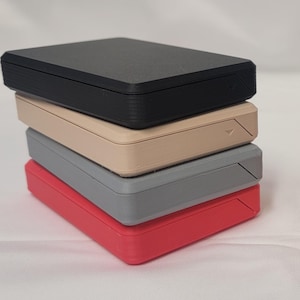 May include: A stack of five rectangular boxes in different colors, including black, beige, gray, red, blue, green, and orange. The boxes are stacked on top of each other, with the black box on top and the orange box on the bottom.