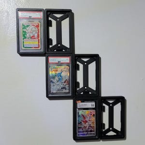 May include: Black plastic card holders displaying graded trading cards. The cards feature colorful illustrations and are encased in clear plastic. The holders are mounted on a white wall.