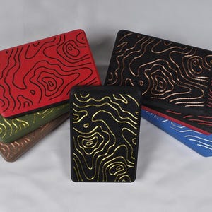 May include: A collection of rectangular, topographic-patterned cases in various colors. The cases feature a raised design with contrasting colors, including red with black, black with gold, and blue with red. Other colors include brown and green.
