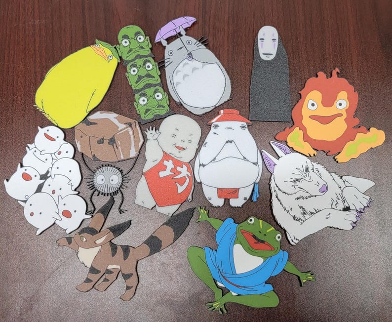 May include: A collection of 14 colorful felt cutouts featuring characters from popular Japanese animated films. The characters include Totoro, No Face, a white wolf, a frog, a red-haired boy, and a group of white and gray creatures.