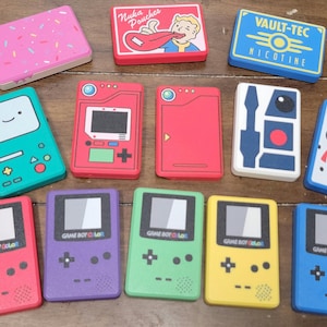 May include: A selection of brightly coloured rectangular cases with diverse designs. Some feature cartoon characters, while others display text such as "Nuka Pouches" and "VAULT-TEC NICOTINE." Several cases resemble retro handheld game consoles, with the text "GAME BOY COLOR" visible.