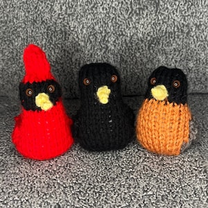 Könnte beinhalten: Drei gestrickte Vogel-Ornamente, ein roter Kardinal, ein schwarzer Vogel und ein orangefarbener Rotkehlchen. Die Vögel haben schwarze Augen und gelbe Schnäbel.