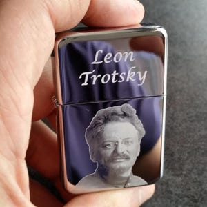 May include: A silver-colored lighter with a purple background. The lighter features the name "Leon Trotsky" in white script and a black and white portrait of a man. The lighter is held by a hand.