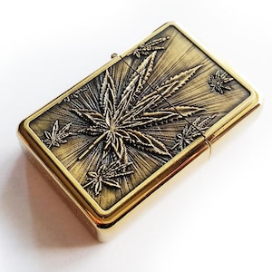 May include: A rectangular, gold-coloured lighter with a detailed cannabis leaf design on the top. The design features a central leaf with radiating lines and smaller leaves around it. The lighter has a shiny, metallic finish.