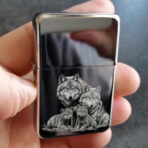 May include: Black lighter with a silver trim, featuring a detailed illustration of a wolf pack. The artwork depicts a family of wolves, with adult wolves and their pups, in shades of grey and white.