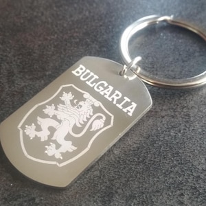 May include: A silver-colored keychain with a rectangular tag. The tag features the word "BULGARIA" in white capital letters above a white emblem of a lion rampant within a shield. The keychain ring is also silver.