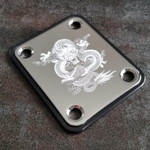 Könnte beinhalten: Eine rechteckige Gitarrenhalsplatte aus Metall mit polierter Chromoberfläche und schwarzem Rand. In der Mitte befindet sich eine detaillierte, weiß lasergravierte Grafik eines gewundenen Drachen. Die Platte hat vier Schraublöcher.