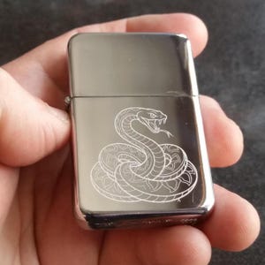 May include: A rectangular, polished chrome metal lighter featuring a detailed, fine-line laser engraving of a coiled snake with its mouth open, showing fangs and a forked tongue. The lighter has a classic flip-top lid and a smooth, reflective surface.