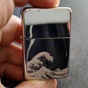 May include: A silver-coloured lighter with a black panel featuring a detailed wave design. The lighter is rectangular and has a shiny, reflective surface. The wave design is in shades of white and grey.