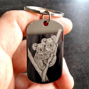 May include: A silver-toned metal keychain featuring a rectangular dog tag pendant with rounded corners. The surface of the pendant is engraved with a detailed, white-toned illustration of two koalas clinging to a tree branch against a dark background.