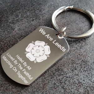 Può includere: Un portachiavi con targhetta argentata con un portachiavi argentato. La targhetta presenta il testo "We Are Leeds" sopra un emblema di rosa bianca. Sotto l'emblema ci sono le parole "Loved By All, Hated By The Faithful, Marching On Together."