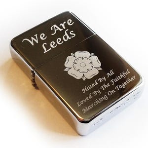 Puede incluir: Un encendedor plateado con las palabras "We Are Leeds" y un emblema floral grabado en la parte delantera. El texto adicional incluye "Hated By All, Loved By The Faithful, Marching On Together". El encendedor tiene una tapa con bisagras y un acabado metálico.